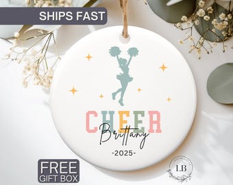 May include: A white ceramic ornament with a cheerleader graphic. The word "CHEER" is in pastel colors, with the name "Brittany" and the year "2025" below. The ornament has a string for hanging and star accents.