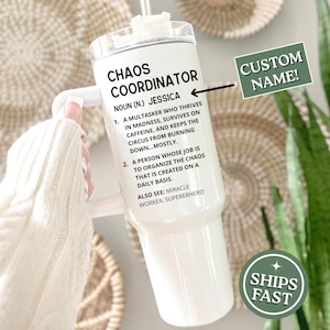 May include: White tumbler with a handle, featuring the text "CHAOS COORDINATOR" and a definition. Includes a space for a custom name. The tumbler is held against a neutral background with woven decor and greenery.