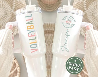 May include: Two white insulated tumblers with lids and handles. One has the word "VOLLEYBALL" in colorful letters. The other has a volleyball graphic, the name "Jennifer", and the number "33". A green "SHIPS FAST" sticker is visible.