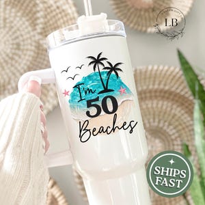 May include: White tumbler with a handle, featuring a beach scene with palm trees, the text "I'm 50 Beaches", and a straw. The design includes a turquoise ocean, sand, and pink starfish. The tumbler is designed for cold beverages.