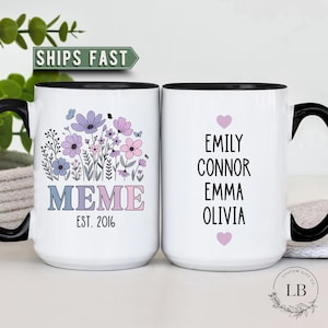 May include: Two white ceramic mugs with black handles and rims. One mug features a floral design with the word "MEME" and "EST. 2016". The other mug has names "EMILY, CONNOR, EMMA, OLIVIA" with pink hearts.