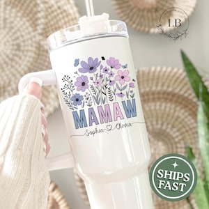 May include: White tumbler with a floral design in purple, blue, and pink. The word "MAMAW" is printed in blue, with "Sophia & Olivia" below. The tumbler has a handle and a straw.