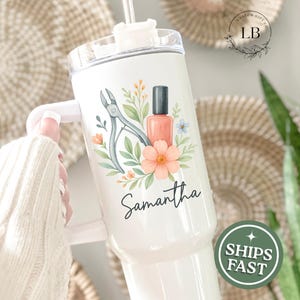 May include: White tumbler with a nail art design. The design includes nail clippers, a bottle of nail polish, and floral accents. The name "Samantha" is written in script. The tumbler has a handle and a straw.