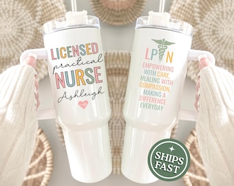 以下が含まれることがあります： ハンドルと透明な蓋が付いた白いステンレス製タンブラー。片面には「LICENSED practical NURSE Ashleigh」という文字とハートのアイコンが描かれています。もう片面には「LPN」とカドゥケウスのシンボル、そしてパステルカラーで「EMPOWERING WITH CARE, HEALING WITH COMPASSION, MAKING A DIFFERENCE EVERYDAY」という文字が記されています。