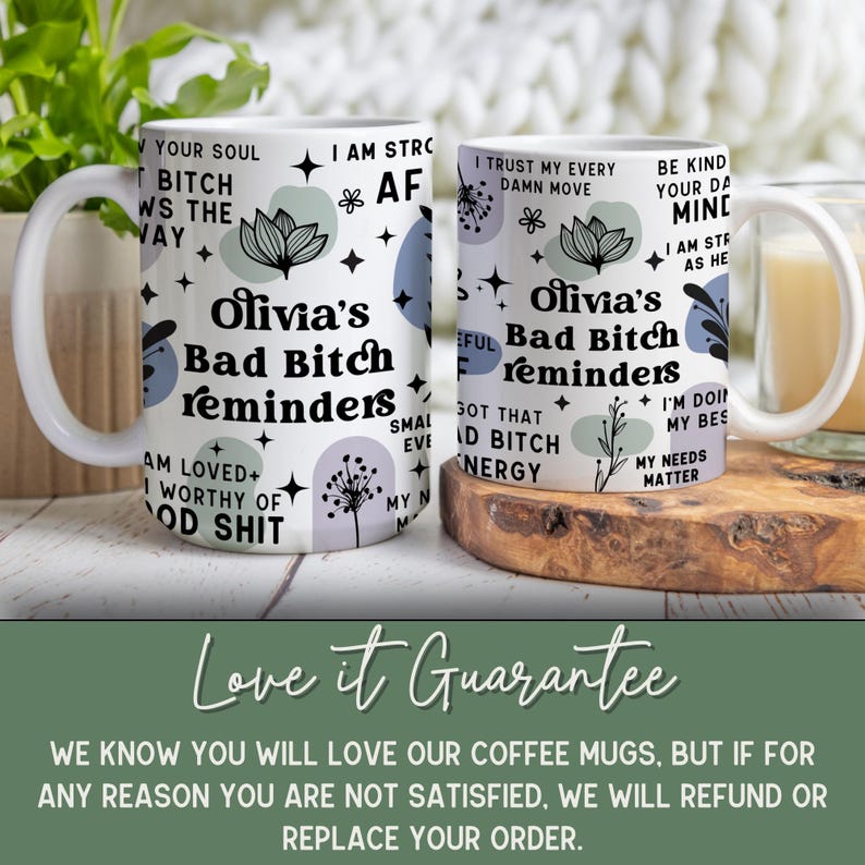 May include: Two white ceramic coffee mugs with the text "Olivia's Bad Bitch Reminders" in black. The mugs have a floral and star design with inspirational phrases. The mugs are on a wooden surface.
