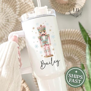 May include: White tumbler featuring a watercolor illustration of a nutcracker soldier in pink, blue, and gold. The name "Bailey" is written in cursive below the image. The tumbler has a white handle and a straw.