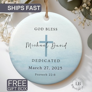 May include: A round, light blue ceramic ornament with a cross and the words "GOD BLESS Michael David DEDICATED March 27, 2025 Proverb 22:6." The ornament has a hole for hanging and is surrounded by greenery.