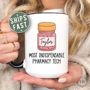 May include: A white ceramic mug with a black handle and rim, featuring a graphic of a pink pill bottle filled with small hearts. The label on the bottle reads Taylor, and the text below the graphic reads MOST INDISPENSABLE PHARMACY TECH.