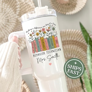 May include: White tumbler with a handle, straw, and lid. The tumbler features the text "Making a difference, one day at a time" and "BEHAVIOR TECHNICIAN Mrs. Smith." Colorful books with inspirational words and floral accents decorate the tumbler.