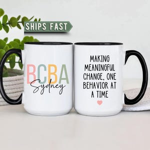 May include: Two white ceramic mugs with black handles and rims. One mug displays "BCBA Sydney" in colorful letters, the other reads "MAKING MEANINGFUL CHANGE, ONE BEHAVIOR AT A TIME" with a pink heart. The mugs are on a white surface.