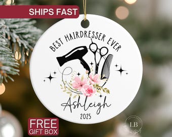 Puede incluir: Adorno redondo de cerámica blanca con el texto "BEST HAIRDRESSER EVER" y el nombre "Ashleigh". El diseño incluye un secador de pelo negro, tijeras, un peine y flores rosas. También se incluye el año 2025.