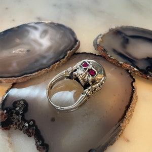 Op de afbeelding: Een zilveren schedelring met rode edelsteenogen. De ring ligt op een grijze en bruine agaatonderzetter.