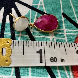 Puede incluir: Un par de pendientes con una piedra rosa en forma de lágrima y una piedra blanca redonda. Los pendientes se miden con una regla, mostrando que tienen un poco más de 2,5 cm de largo.