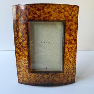 May include: A rectangular picture frame featuring a glossy, mottled amber and dark brown tortoiseshell pattern. The frame has a curved top and bottom edge, a smooth finish, and a central rectangular opening with a light gray backing insert.