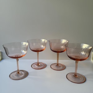 May include: Four pink champagne coupe glasses with a spiral design on the bowl. The glasses have a stem and a round base. The glasses are arranged in a row on a white surface.
