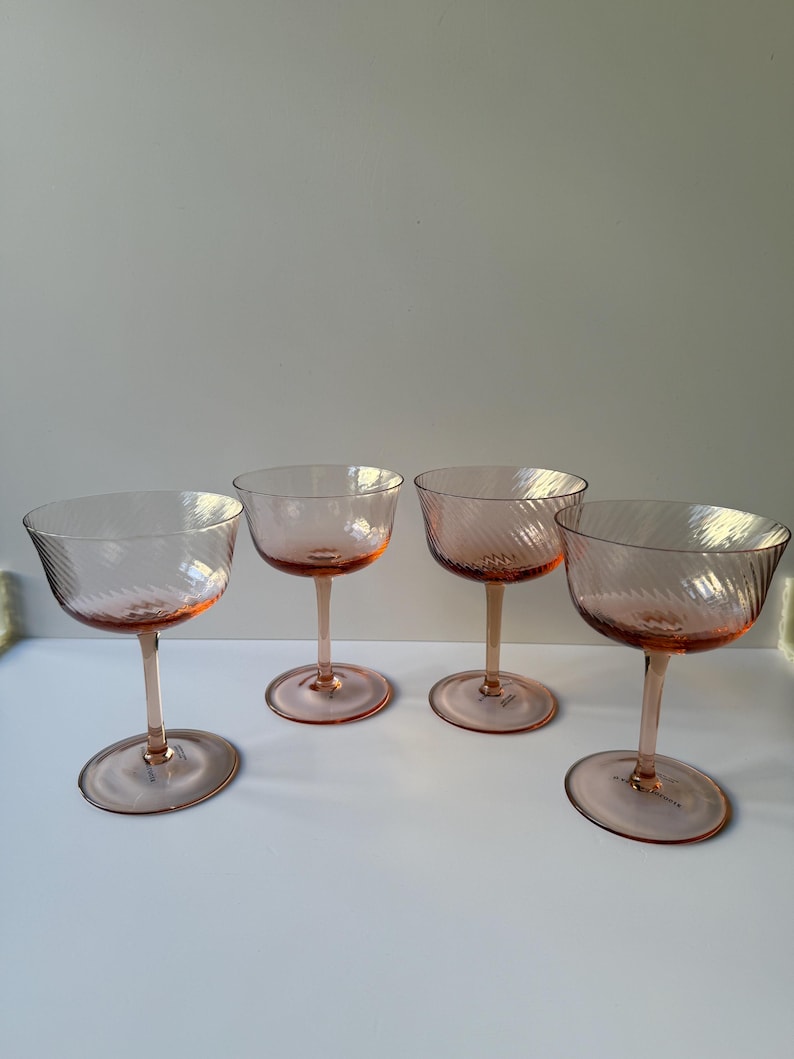 May include: Four pink champagne coupe glasses with a spiral design on the bowl. The glasses have a stem and a round base. The glasses are arranged in a row on a white surface.
