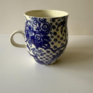 May include: A ceramic mug with a cream-colored interior and a white handle. The exterior features a blue floral pattern. The mug has a wide, rounded shape and is suitable for hot beverages. The design is reminiscent of classic blue and white china.