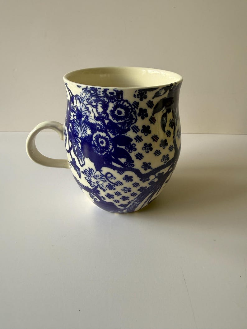 May include: A ceramic mug with a cream-colored interior and a white handle. The exterior features a blue floral pattern. The mug has a wide, rounded shape and is suitable for hot beverages. The design is reminiscent of classic blue and white china.