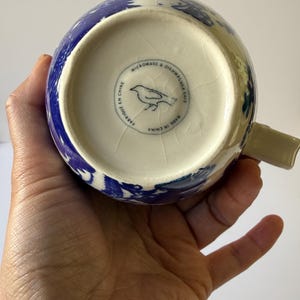 May include: A blue and white ceramic mug with a floral design. The bottom of the mug has a circular stamp with a bird illustration and the text "MICROWAVE & DISHWASHER SAFE", "FABRIQUE EN CHINE", and "MADE IN CHINA".