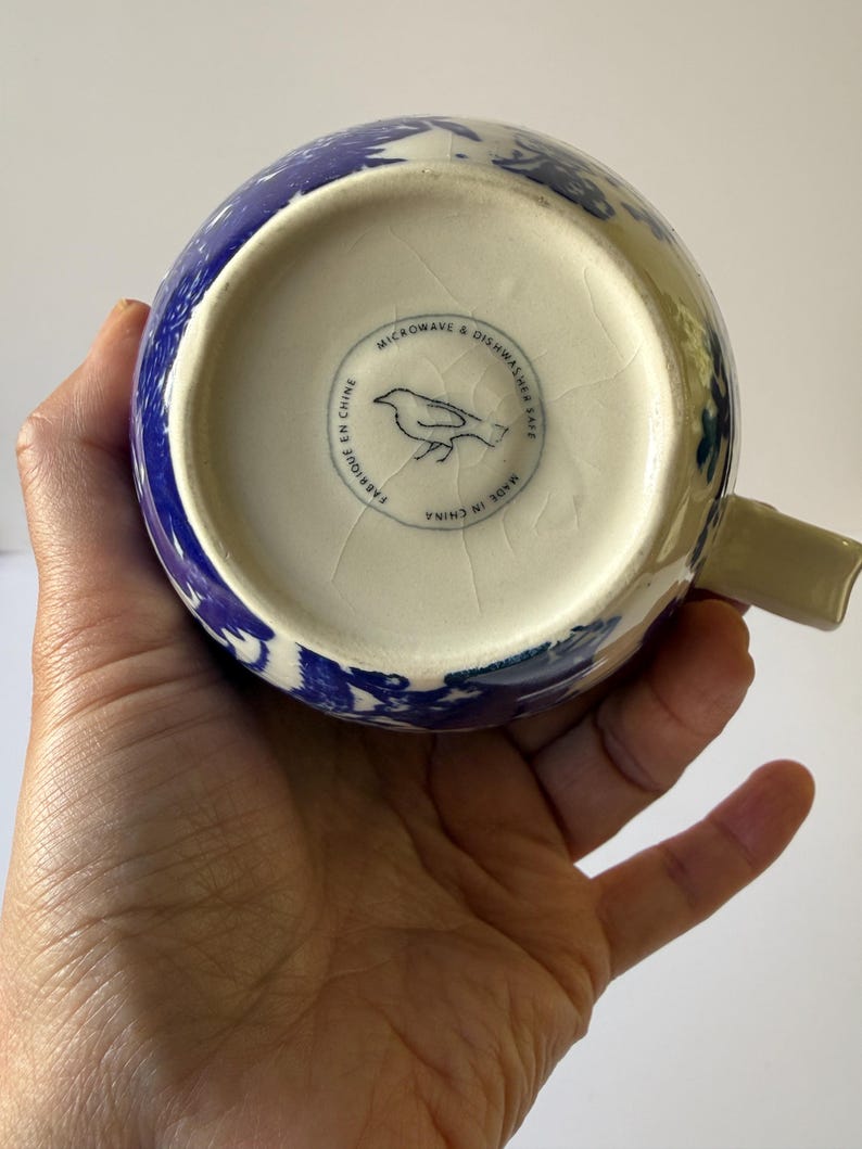 May include: A blue and white ceramic mug with a floral design. The bottom of the mug has a circular stamp with a bird illustration and the text "MICROWAVE & DISHWASHER SAFE", "FABRIQUE EN CHINE", and "MADE IN CHINA".
