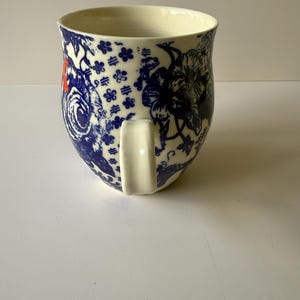 May include: A ceramic mug with a cream-colored interior and a white handle. The exterior features a blue and white floral pattern with red accents. The mug has a wide opening and a rounded base, suitable for hot beverages.