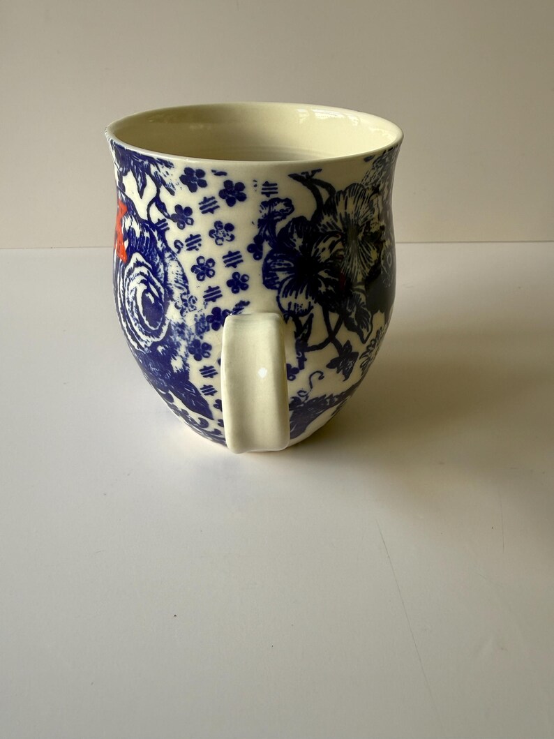 May include: A ceramic mug with a cream-colored interior and a white handle. The exterior features a blue and white floral pattern with red accents. The mug has a wide opening and a rounded base, suitable for hot beverages.