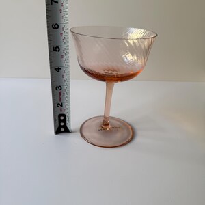 May include: A pink-tinted coupe glass with a spiral design. The glass is approximately 6 inches tall. The stem and base are also pink. The glass is on a white surface.