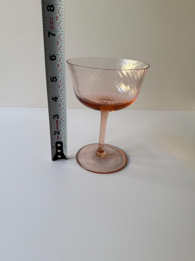 May include: A pink-tinted coupe glass with a spiral design. The glass is approximately 6 inches tall. The stem and base are also pink. The glass is on a white surface.