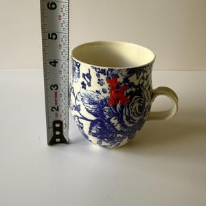 May include: A ceramic mug with a blue floral pattern and a red letter "K". The mug is approximately 4 inches tall. The handle and interior are cream-colored.