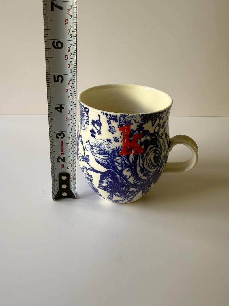 May include: A ceramic mug with a blue floral pattern and a red letter "K". The mug is approximately 4 inches tall. The handle and interior are cream-colored.