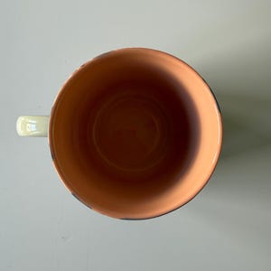 Puede incluir: Vista cenital de una taza de cer&aacute;mica con un asa verde claro. El interior de la taza es de color naranja c&aacute;lido y el borde tiene un ribete azul oscuro. La taza est&aacute; dise&ntilde;ada para bebidas.