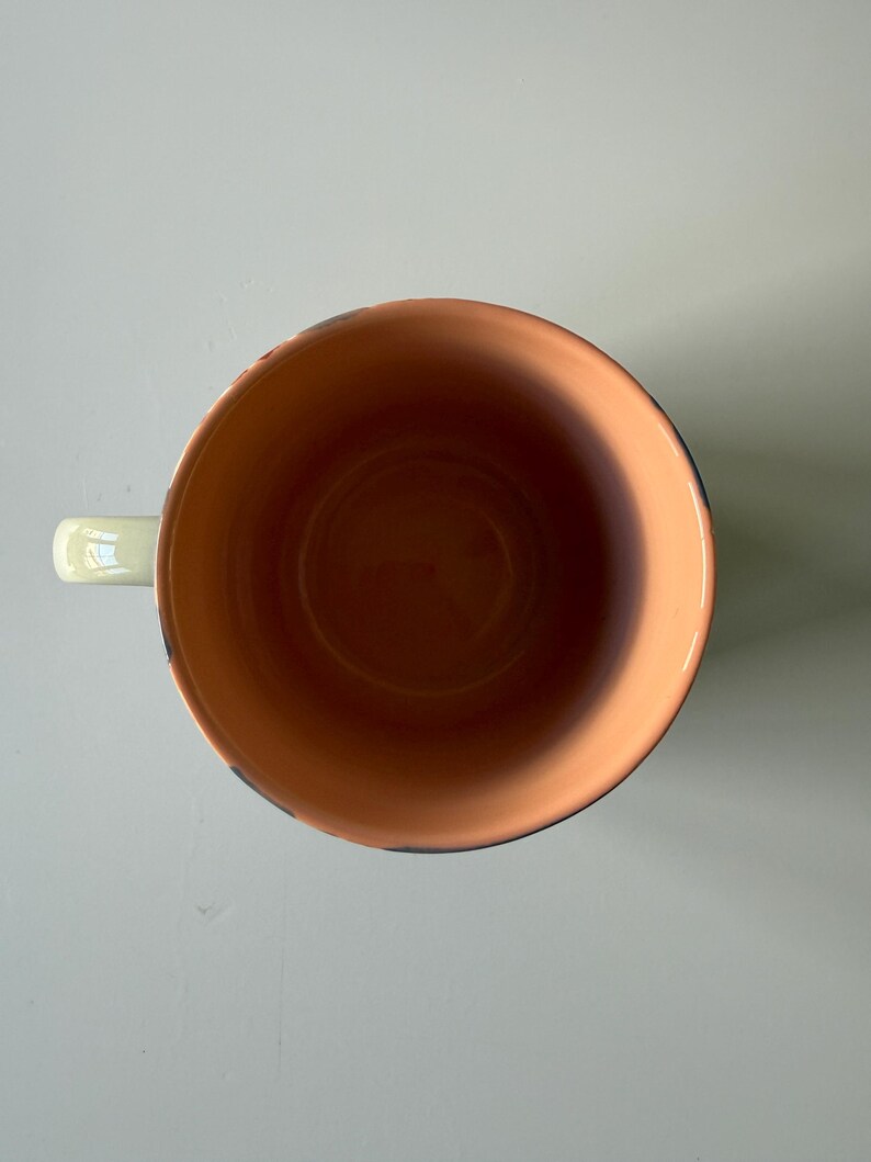 Puede incluir: Vista cenital de una taza de cer&aacute;mica con un asa verde claro. El interior de la taza es de color naranja c&aacute;lido y el borde tiene un ribete azul oscuro. La taza est&aacute; dise&ntilde;ada para bebidas.