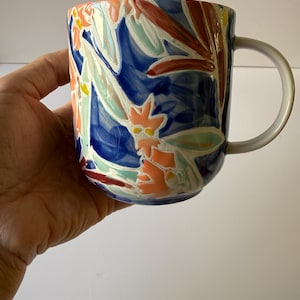Puede incluir: Una taza de cer&aacute;mica con un asa blanca y un dise&ntilde;o floral vibrante. La taza presenta un fondo azul oscuro con flores coloridas en tonos naranja, rojo y verde claro. La taza se sostiene en una mano.