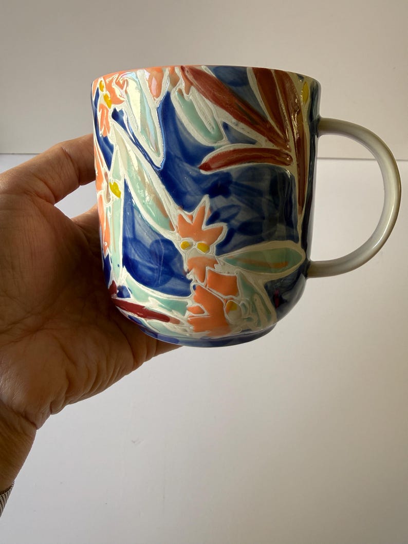 Puede incluir: Una taza de cer&aacute;mica con un asa blanca y un dise&ntilde;o floral vibrante. La taza presenta un fondo azul oscuro con flores coloridas en tonos naranja, rojo y verde claro. La taza se sostiene en una mano.