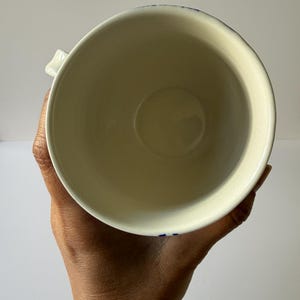 May include: A cream-colored ceramic mug with a blue floral design around the rim. The mug is held in a person's hand, showcasing its interior and handle. The mug has a simple, classic design.
