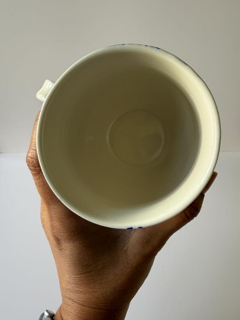 May include: A cream-colored ceramic mug with a blue floral design around the rim. The mug is held in a person's hand, showcasing its interior and handle. The mug has a simple, classic design.