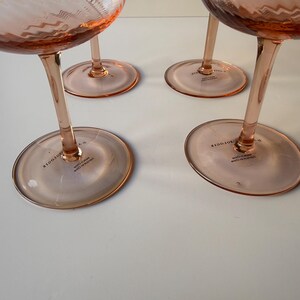 May include: Four pink-tinted stemmed glasses with a textured bowl and a slender stem. The glasses have a wide, circular base. The text "BODOTE" is printed on the base of each glass. The glasses are arranged on a white surface.