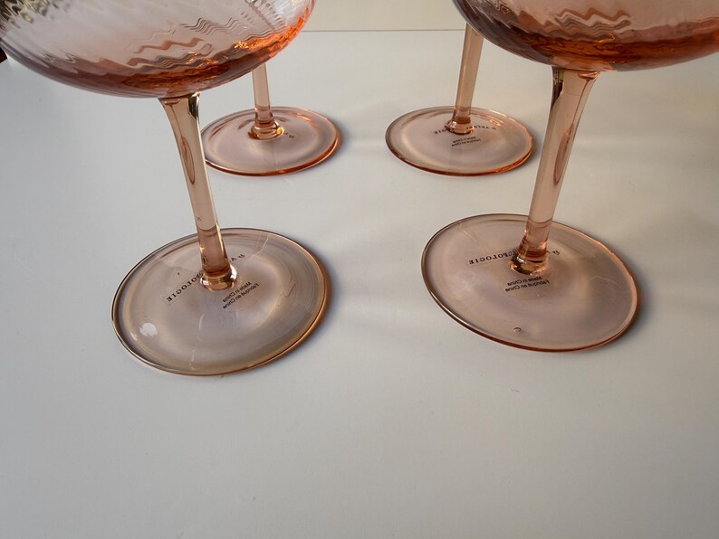 May include: Four pink-tinted stemmed glasses with a textured bowl and a slender stem. The glasses have a wide, circular base. The text "BODOTE" is printed on the base of each glass. The glasses are arranged on a white surface.