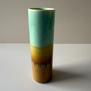 May include: A tall, cylindrical ceramic vase with a smooth, glossy finish. The top half is a vibrant turquoise, transitioning to a warm yellow and then a rich brown at the base. The vase has a simple, elegant design.