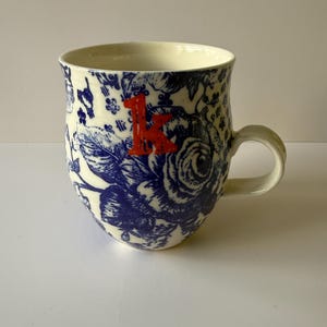 May include: A ceramic mug with a white background and a blue floral pattern. The mug has a red letter "K" on the front. The handle is white and the mug is approximately 4 inches tall.