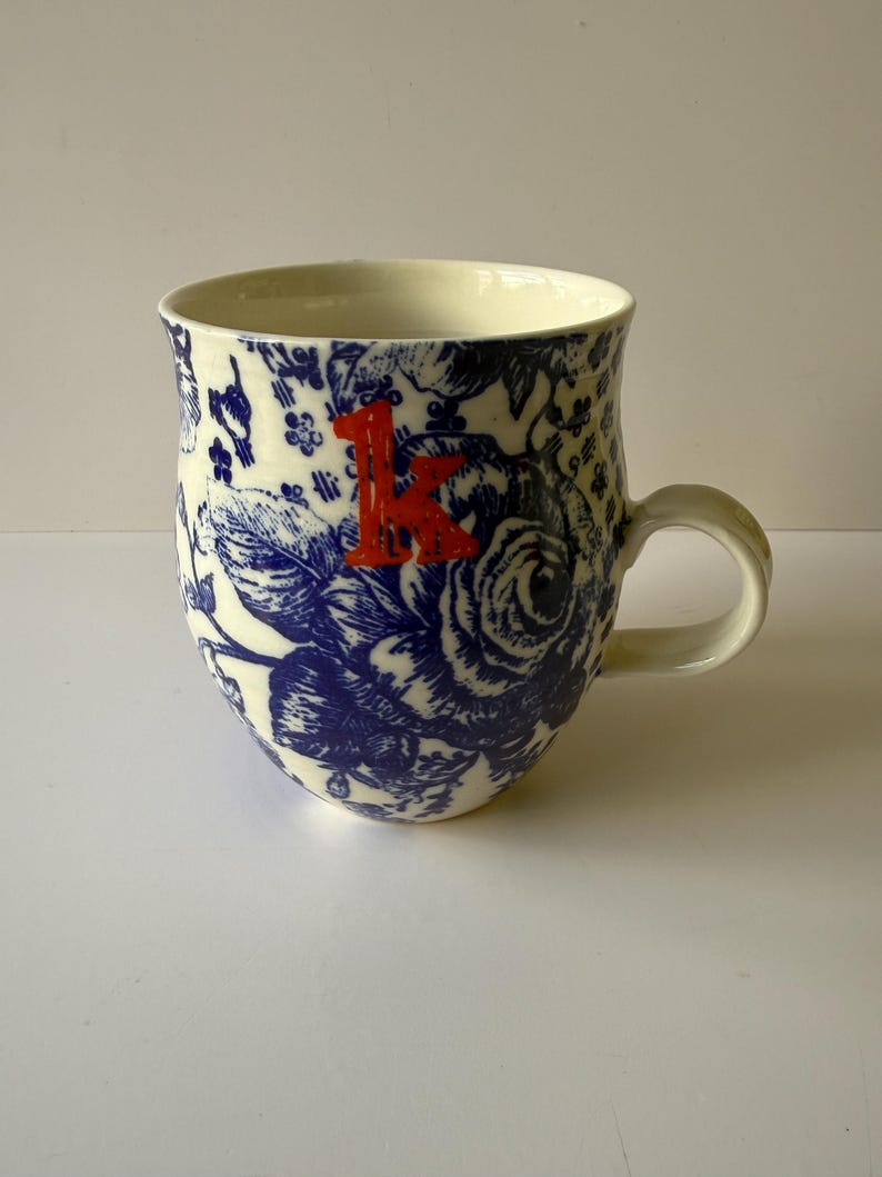 May include: A ceramic mug with a white background and a blue floral pattern. The mug has a red letter "K" on the front. The handle is white and the mug is approximately 4 inches tall.