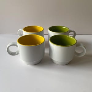 May include: Four white ceramic mugs with a rounded handle. Two mugs have a bright yellow interior, and two have a green interior. The mugs are stacked and have a simple, modern design.