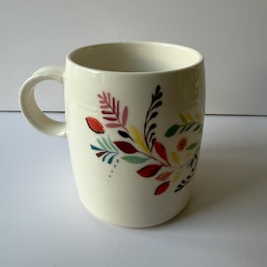 May include: Cream-colored ceramic mug with a curved handle. The mug is decorated with a colorful floral design featuring red, yellow, and green leaves. The mug is a standard size for coffee or tea.