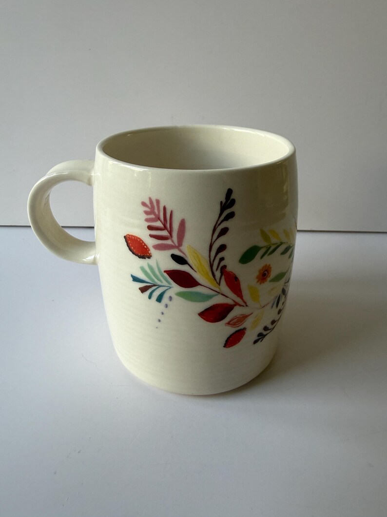 May include: Cream-colored ceramic mug with a curved handle. The mug is decorated with a colorful floral design featuring red, yellow, and green leaves. The mug is a standard size for coffee or tea.