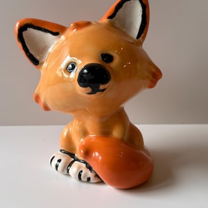 May include: A ceramic fox figurine in a sitting position. The fox is primarily orange with black and white accents on the ears and feet. It has a glossy finish and a black nose and eyes. The tail is curled up.