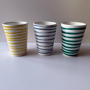 May include: Three white ceramic tumblers with horizontal stripes in yellow, blue, and green. The tumblers have a simple, cylindrical shape and a white interior. The stripes are evenly spaced and add a decorative element to the drinkware.