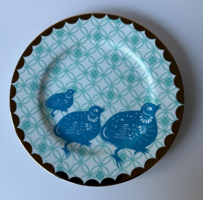 May include: A decorative plate with a teal and white geometric pattern and a scalloped brown rim. Three blue bird illustrations are featured on the plate. The plate is a stylish addition to any home decor.