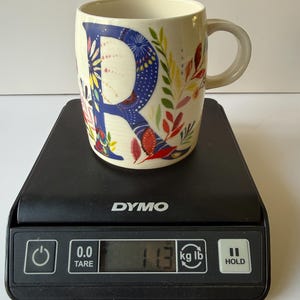 May include: Cream-colored ceramic mug with a large blue letter "R" decorated with colorful floral patterns. The mug is on a black digital scale, displaying a weight of 11.3 kg.