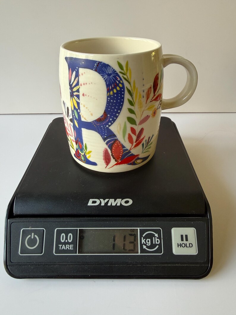 May include: Cream-colored ceramic mug with a large blue letter "R" decorated with colorful floral patterns. The mug is on a black digital scale, displaying a weight of 11.3 kg.