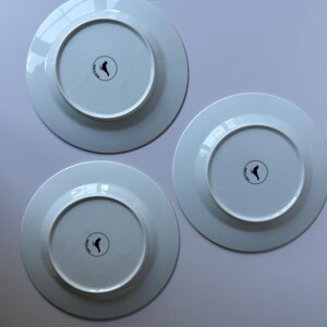 May include: Three white ceramic plates, viewed from the underside, arranged on a light surface. Each plate has a circular indentation in the center with a black logo. The plates are clean and appear unused.